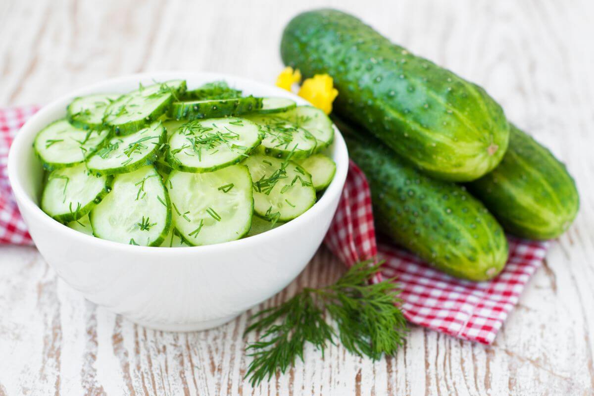 A white bowl of cucumber slices garnished with dill sits on a red checkered cloth, beside three whole cucumbers and a sprig of dill on rustic wood.