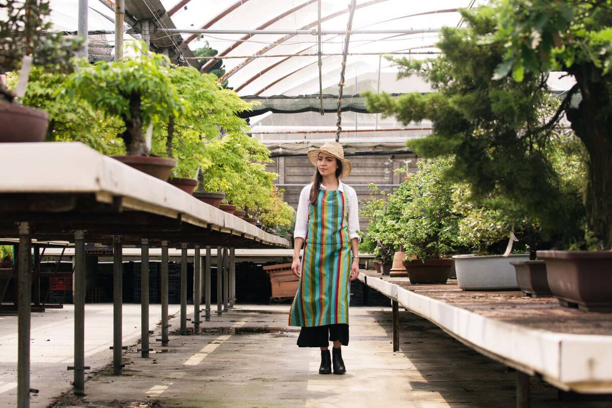 A woman wearing a striped apron and straw hat walks through a greenhouse filled with various potted bonsai trees.