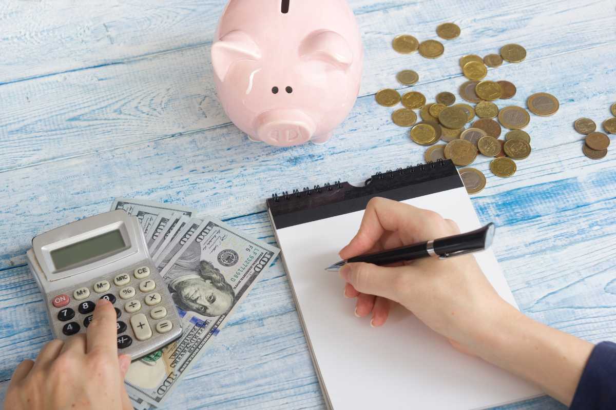 A person is using a calculator while holding a pen to write on a notepad. Near the hand is a stack of U.S. dollar bills, perhaps calculating the cost of hydroponics vs aquaponics. 