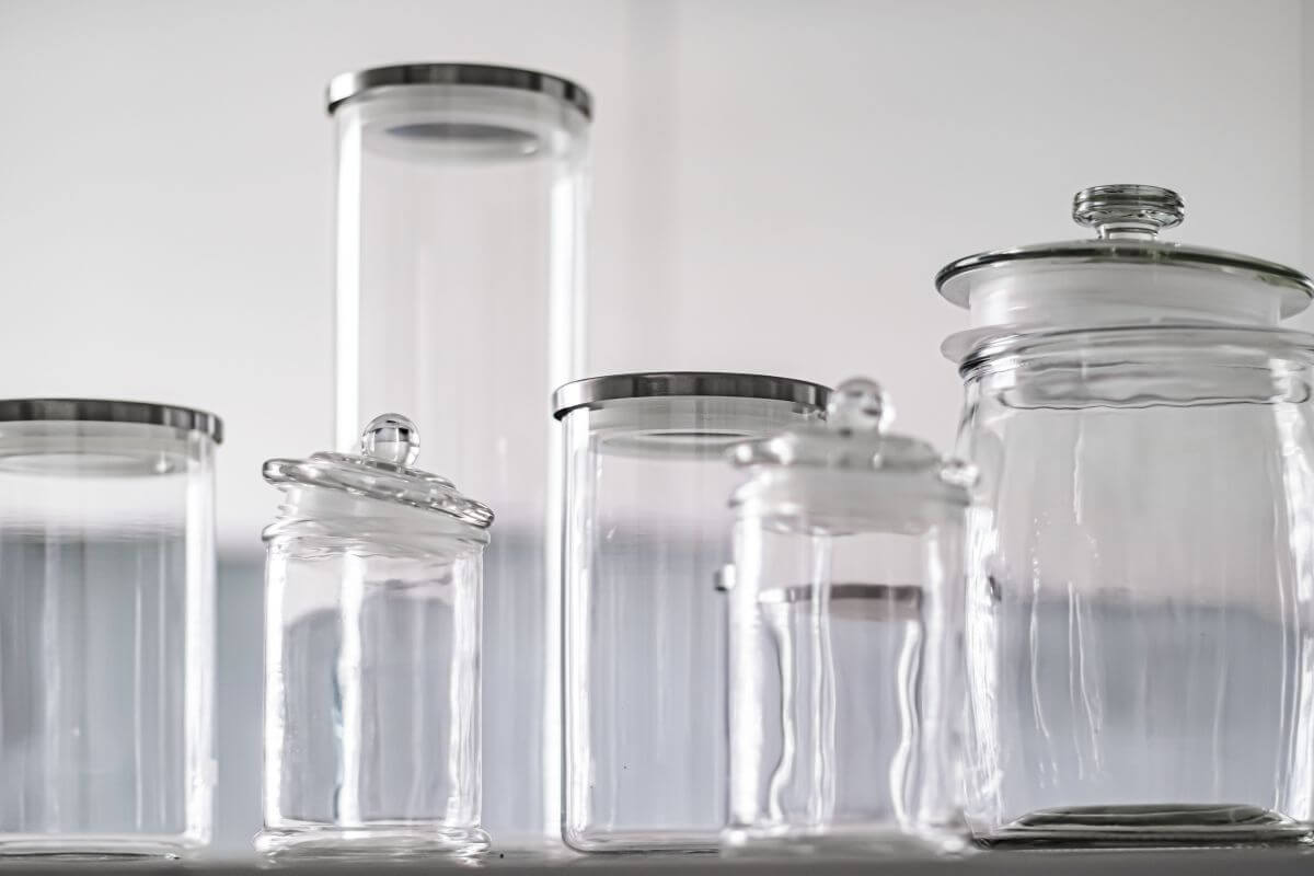 A set of six empty glass jars with metal and glass lids are arranged on a white surface against a soft, blurred background. The jars vary in size and shape, maintaining a cylindrical form and clean, minimalist design.