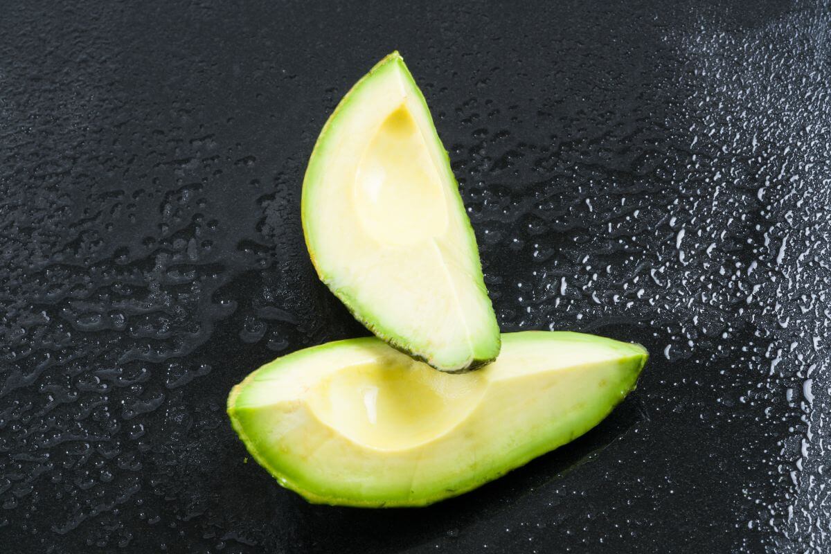 Two slices of hydroponic avocado placed on a dark, textured surface with water droplets visible.