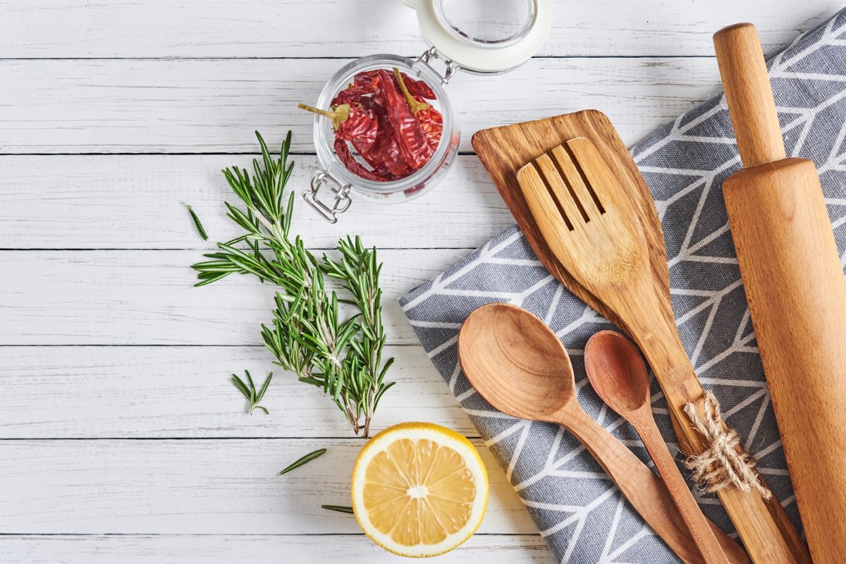 A flat lay image on a white wooden surface showing kitchen utensils including a wooden spatula, spoon, and fork tied with twine, a rolling pin, a glass jar with dried red peppers, a sprig of rosemary, half a lemon, and a gray patterned cloth.