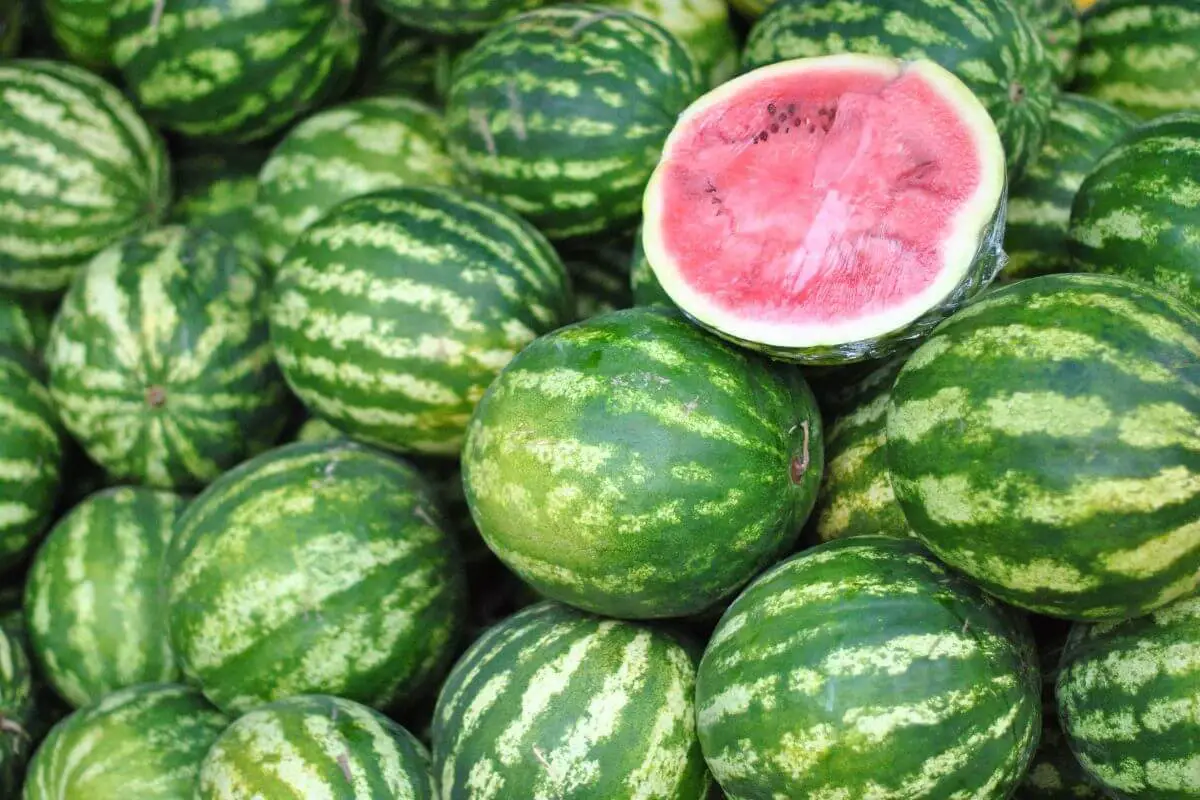 A pile of whole watermelons with green striped rinds. Among them, one watermelon is cut in half, revealing its vibrant red flesh and black seeds. 