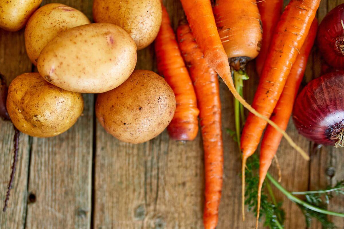 A rustic wooden table with a collection of fresh root vegetables, including several brown potatoes, vibrant orange carrots with green tops, and a few red onions.