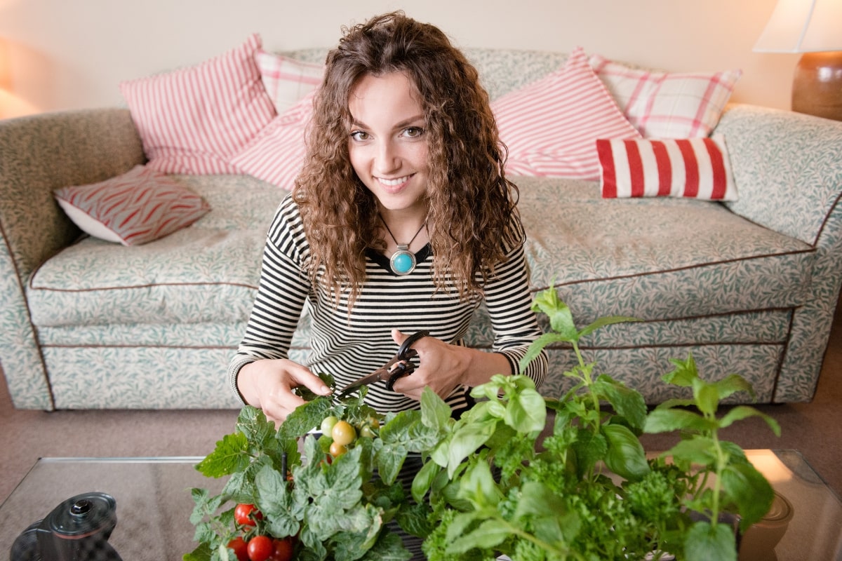 A smiling woman with curly hair wearing a striped top uses scissors to trim fresh herbs from potted plants on a glass table.
