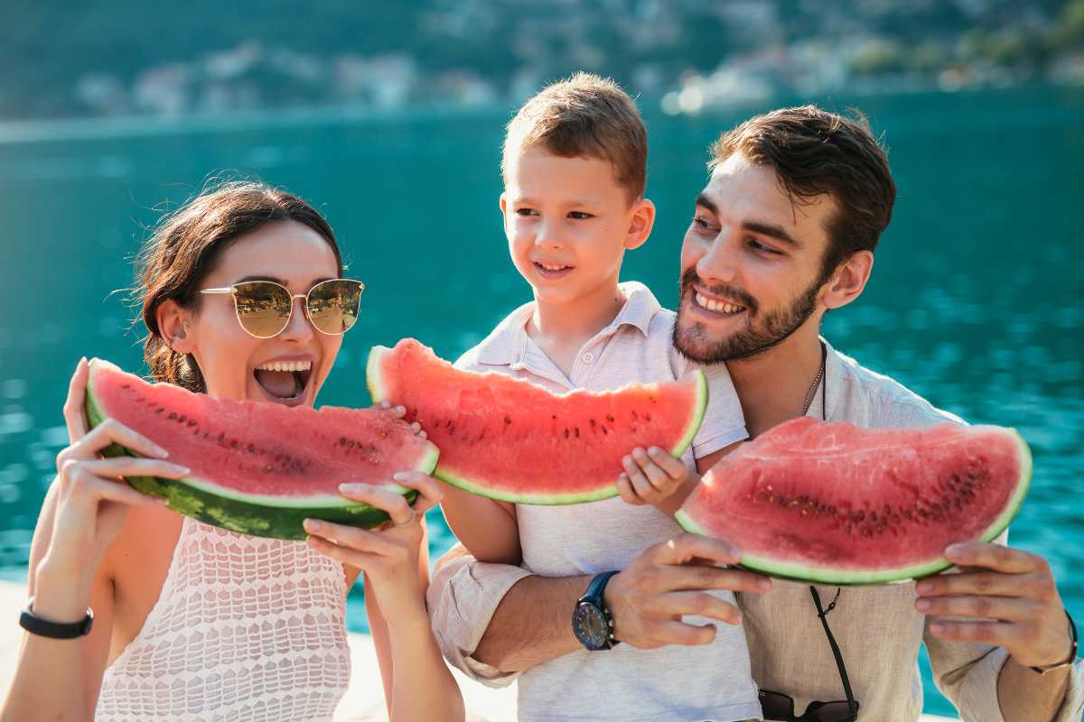 A woman, a man, and a child are enjoying large slices of watermelon by a body of water with a scenic background. 