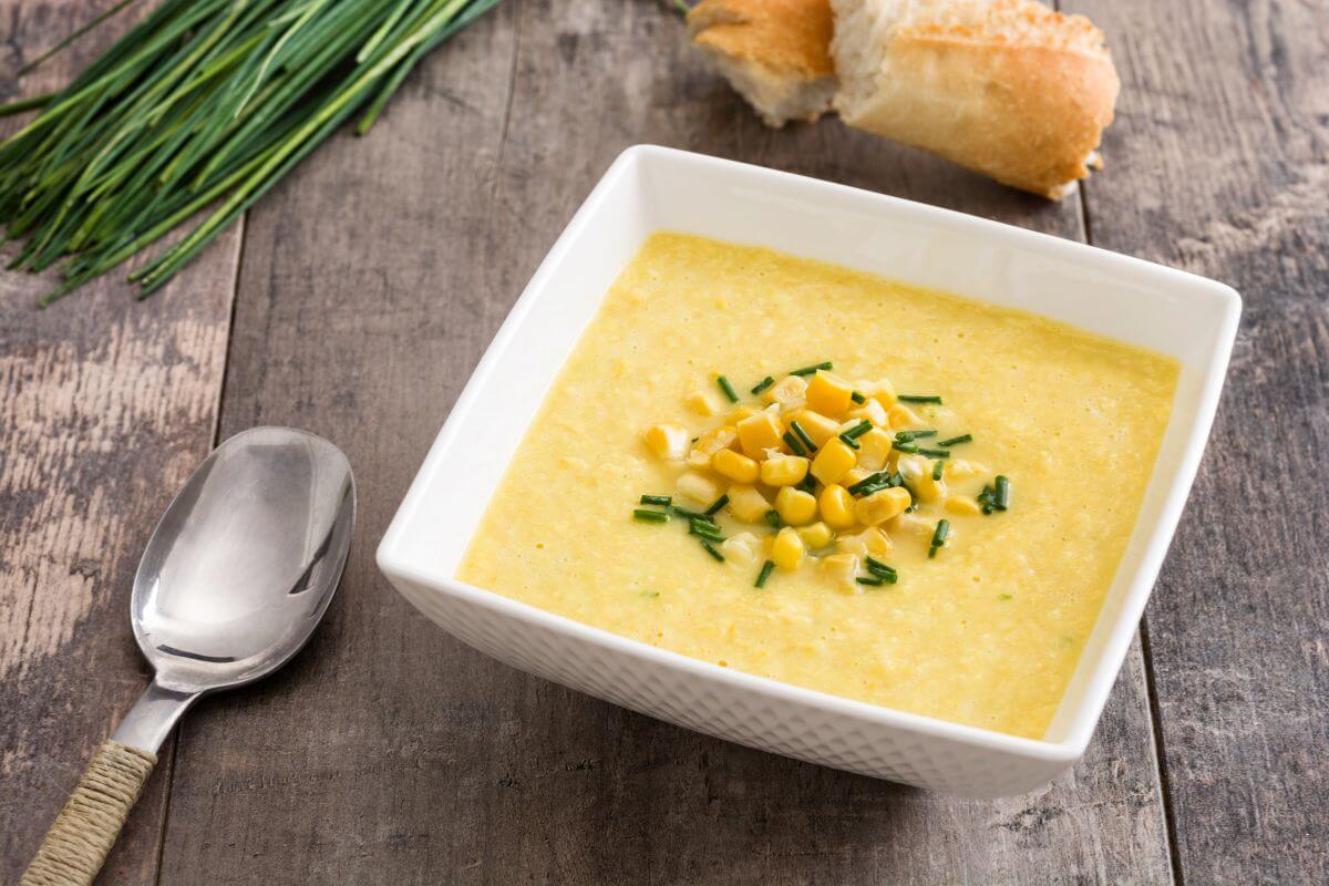 A square white bowl filled with creamy corn soup, garnished with corn kernels and chopped chives. A piece of crusty bread, fresh chives, and a spoon are in the background on a rustic wooden surface.