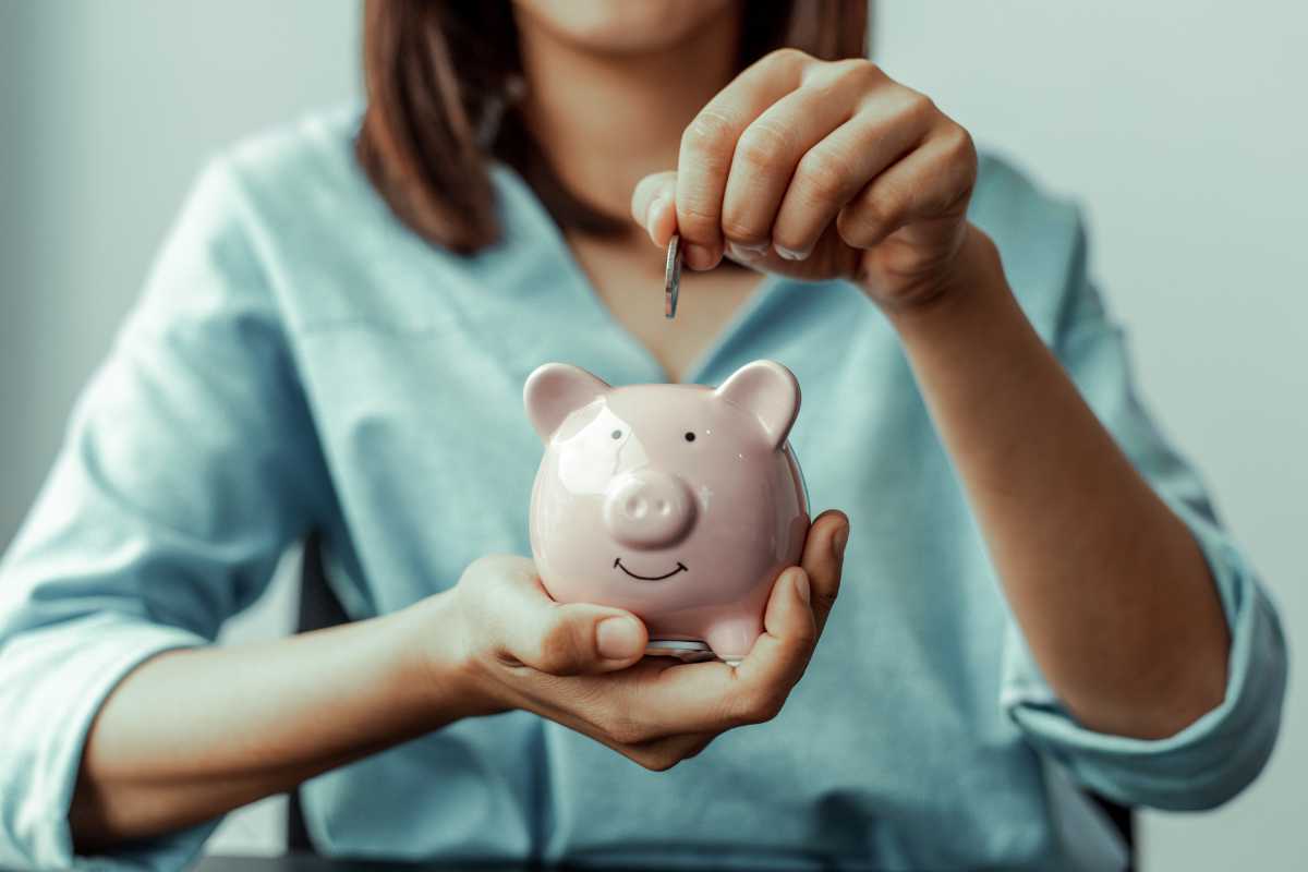 A person wearing a light blue shirt is placing a coin into a pink piggy bank. The piggy bank, with its smiling face, is held in the person's other hand. 