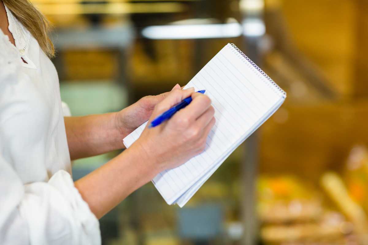 A person in a white shirt is holding an open notepad and writing in it with a blue pen, jotting down composting log. 