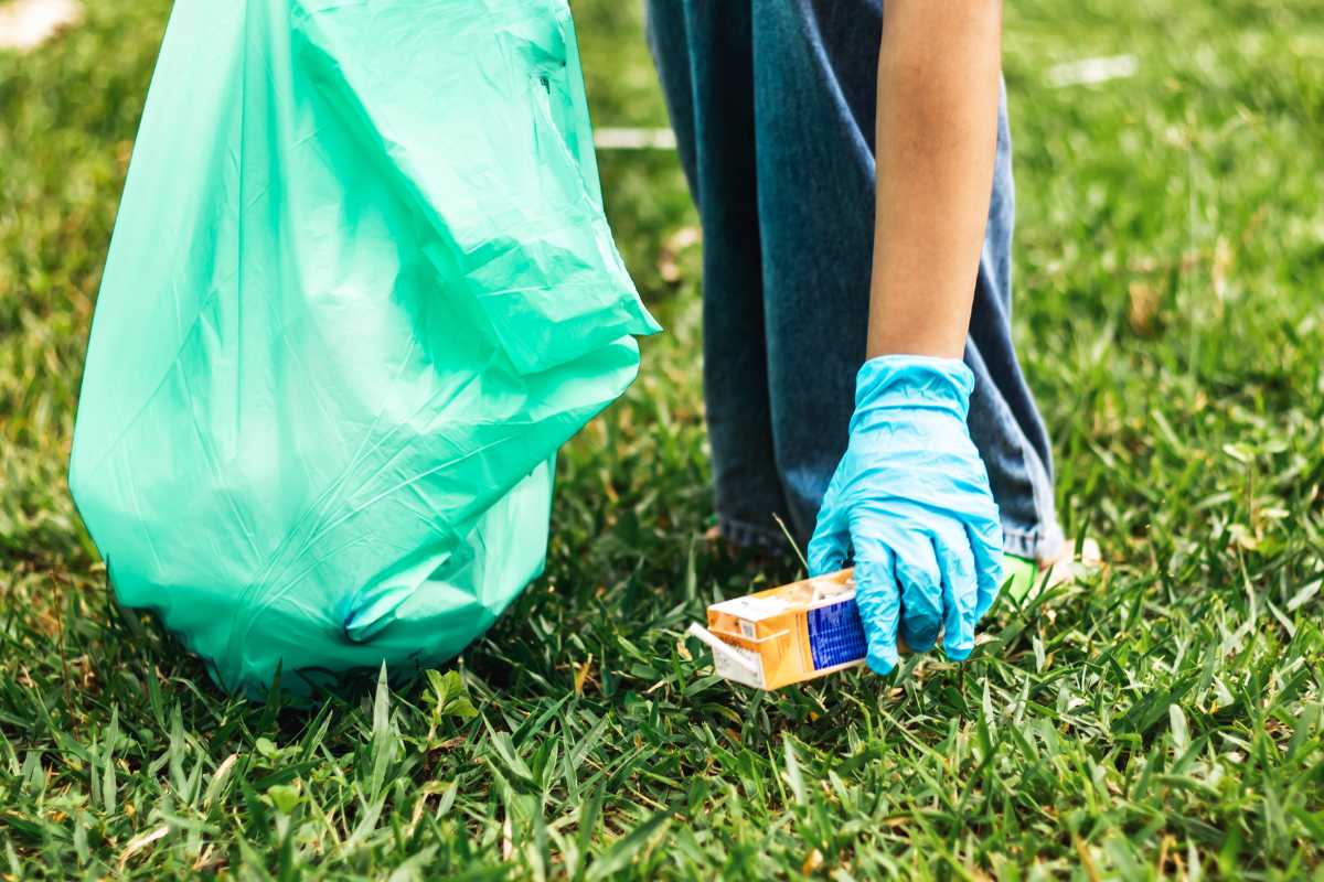 A person wearing blue gloves picks up a trash from the grass and holds a green trash bag in their other hand.