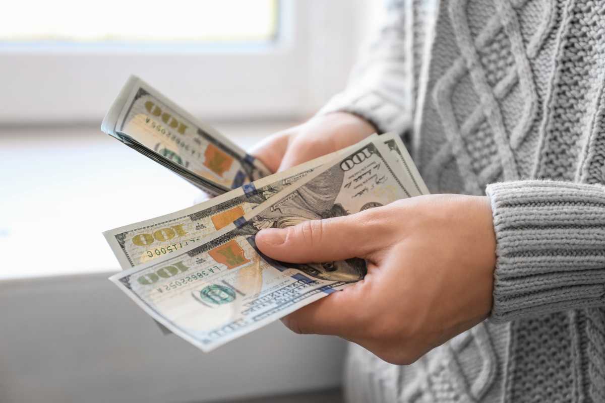 A person wearing a light gray knit sweater is holding a stack of U.S. 100-dollar bills in their hands, counting the money near a window.