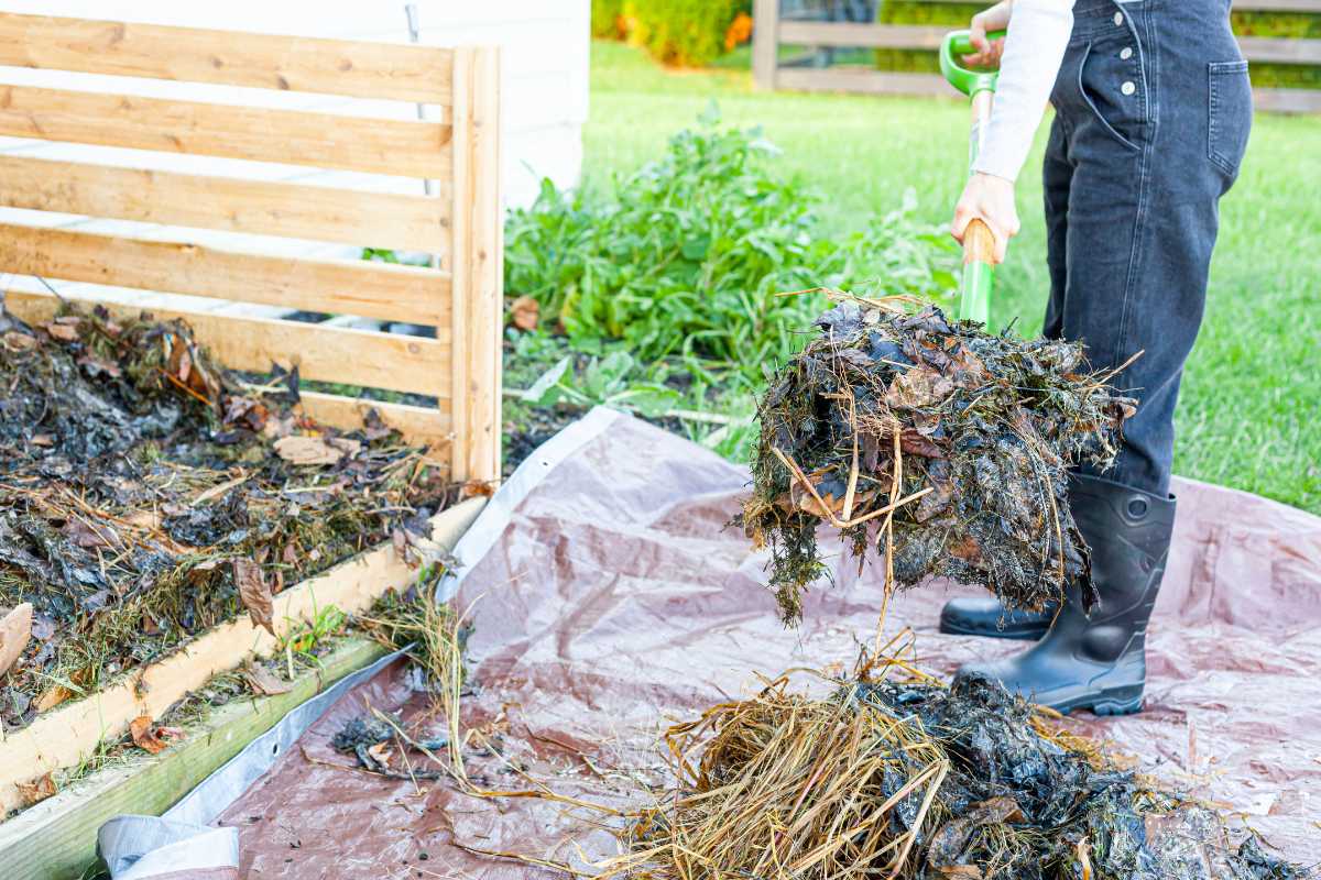 A person wearing black pants and black boots is using a garden tool to turn a pile of leaves, grass, and other compost ingredients on a brown tarp near a wooden compost bin in a backyard.