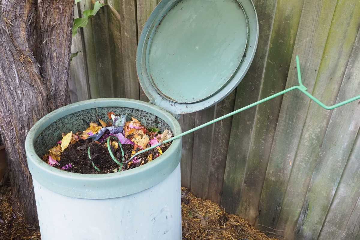 A light blue compost bin with a lid leaning open against a wooden fence contains various compostable materials, including fruit peels, eggshells, and garden waste.