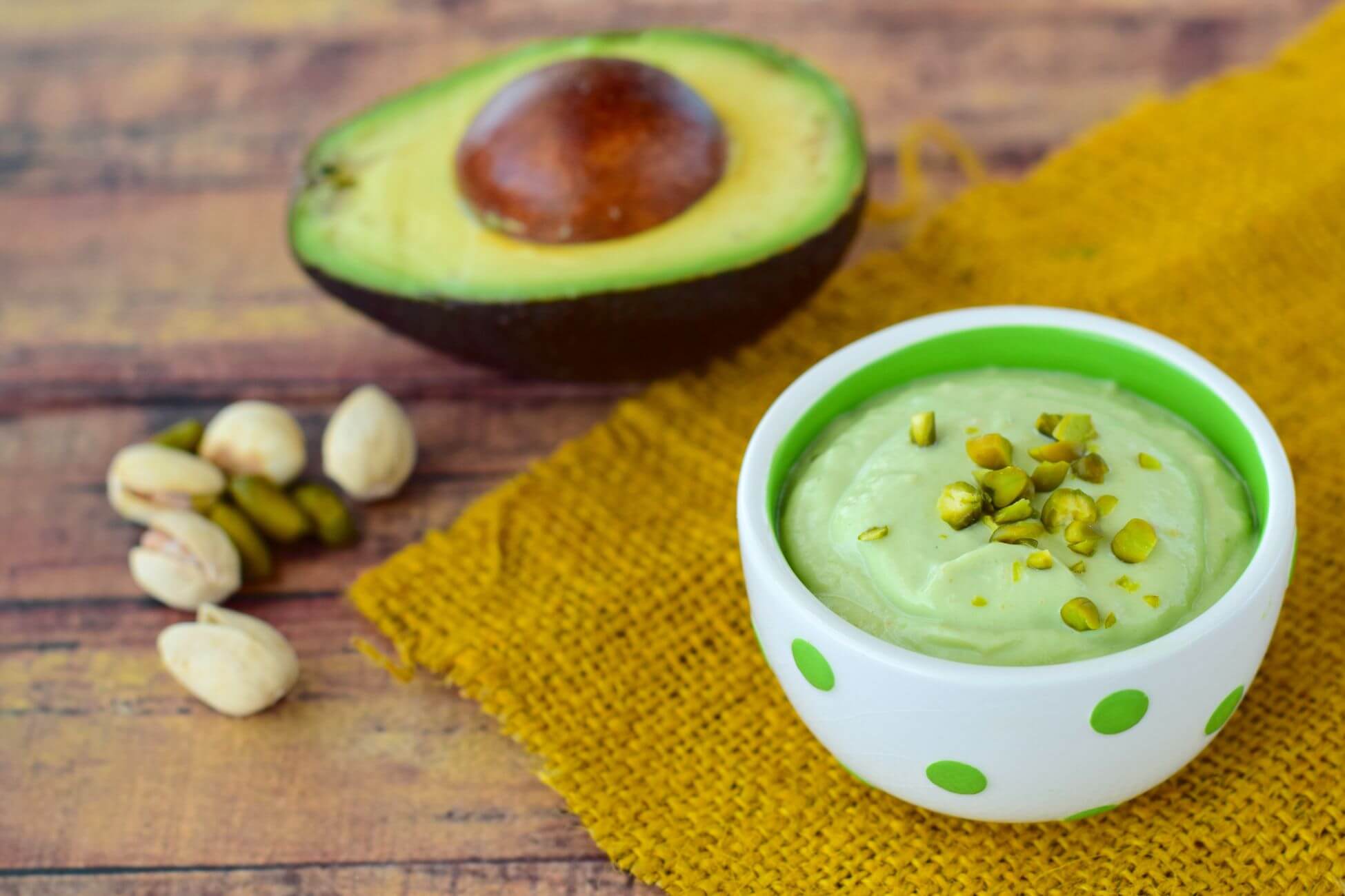 A bowl of avocado pudding topped with chopped pistachios sits on a yellow burlap cloth. 