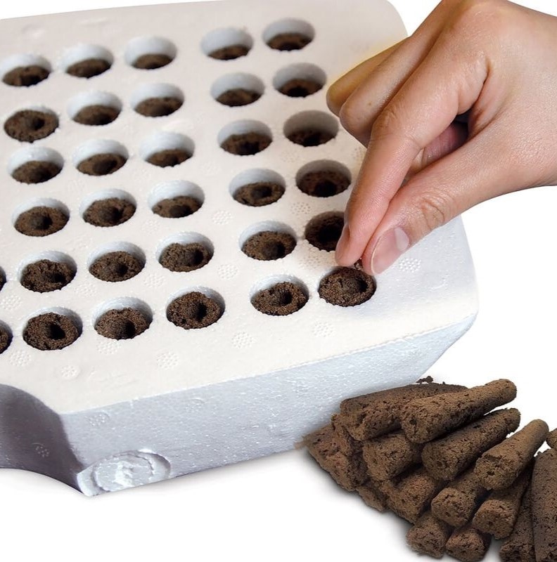 A hand planting seeds or small plants into rows of holes filled with soil in a white tray. The tray, often used in aerogarden setups, has multiple rows and columns of small holes, each containing grow sponges. 