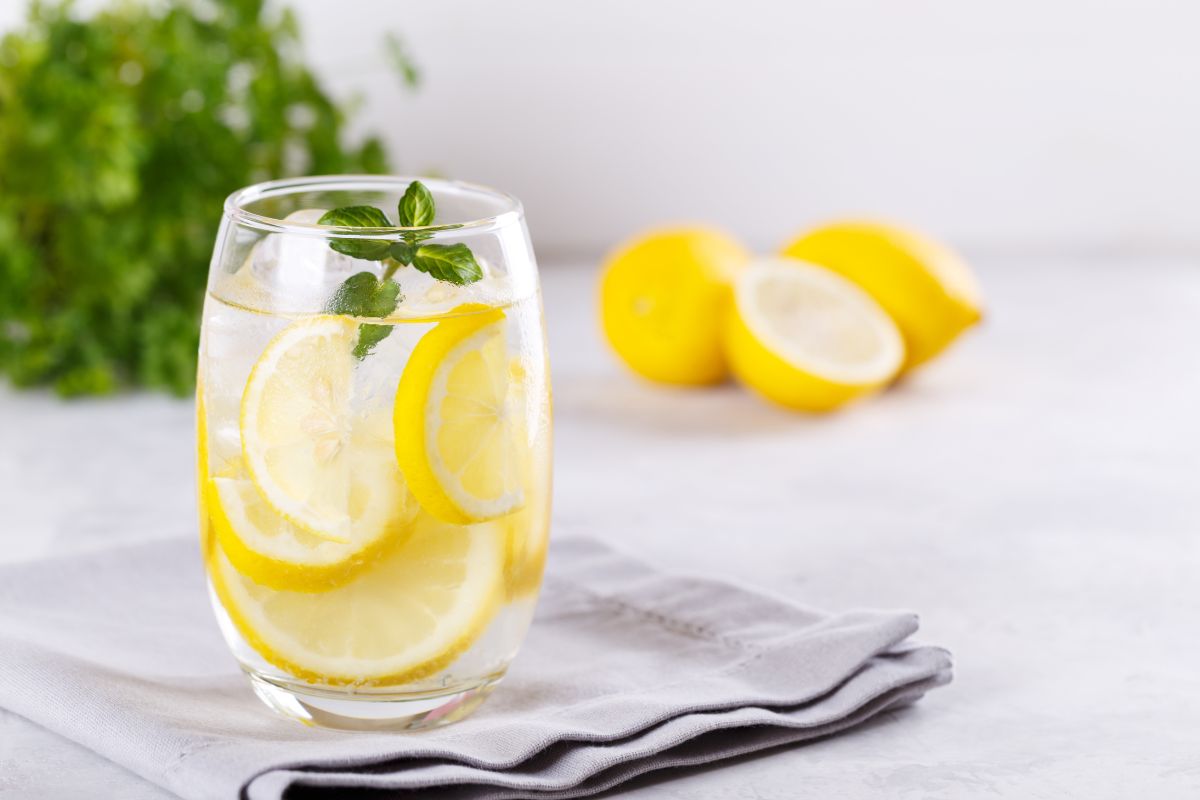 A glass of lemon water with lemon slices and mint on a gray napkin. In the background, two whole lemons, a half lemon, and a blurred leafy green plant on a light gray surface.