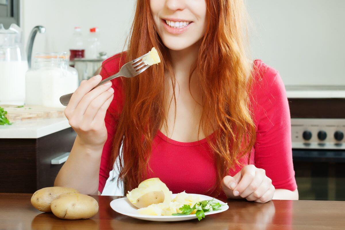 A woman with long red hair, wearing a red top, is holding a fork with a piece of potato close to her mouth. 