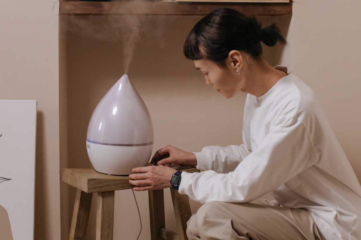 A person in a white long-sleeve shirt is adjusting a white, teardrop-shaped humidifier for grow room use, emitting mist and placed on a small wooden stool.