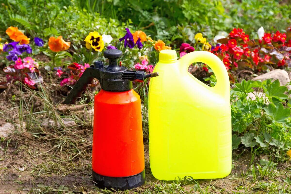 An orange garden sprayer with a black nozzle and a yellow plastic container of liquid fertilizer with a handle, placed on the grass.