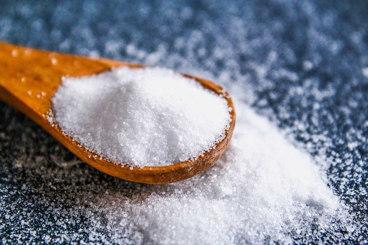 A wooden spoon holding a heap of granulated white sugar, with some sugar scattered on a dark, textured surface. 