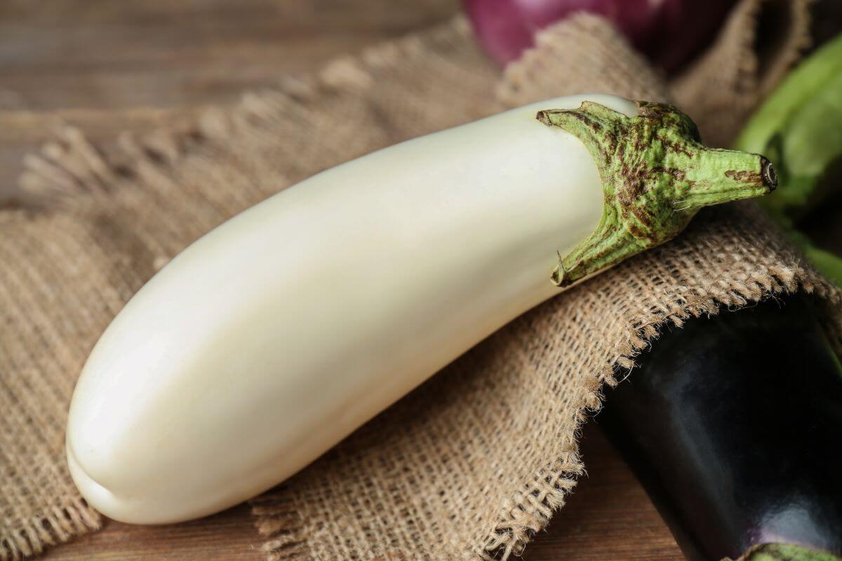 A close-up of a white eggplant with a green stem on a burlap cloth, with other vegetables slightly out of focus in the background, including a hint of a dark purple eggplant. 