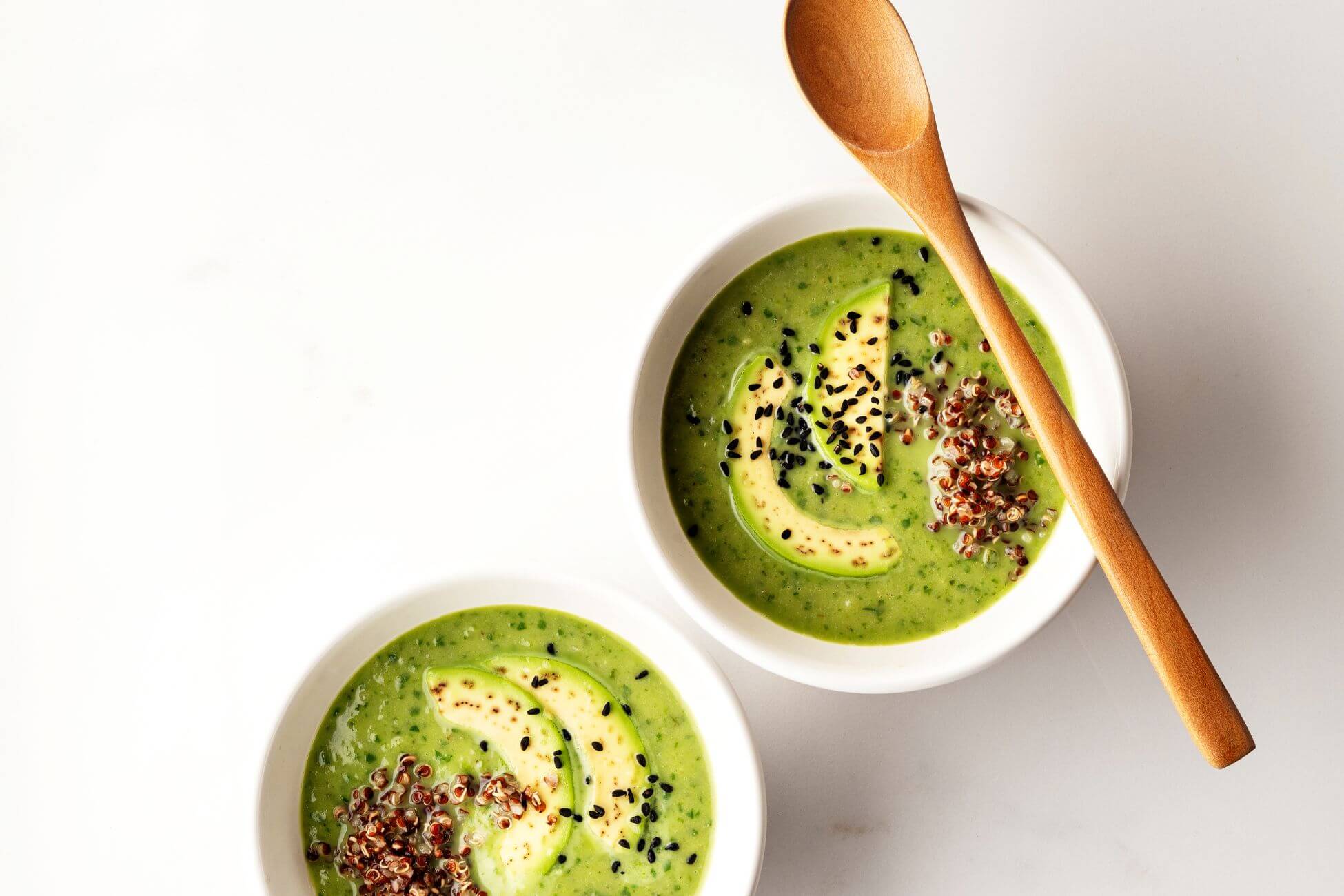 Two white bowls of green avocado soup garnished with avocado slices, quinoa, and black sesame seeds sit on a white surface. 