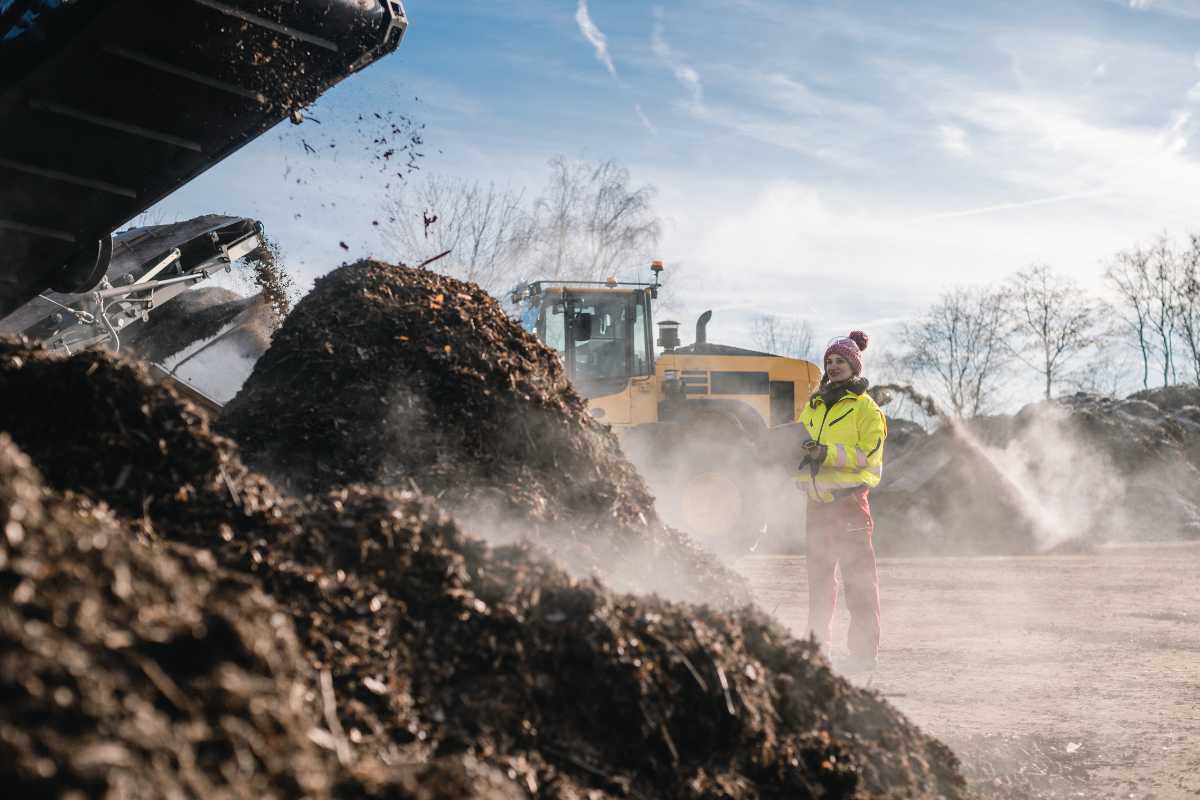 A person in a high-visibility jacket and red pants stands near steaming piles of compost being processed by heavy machinery on a clear day. 