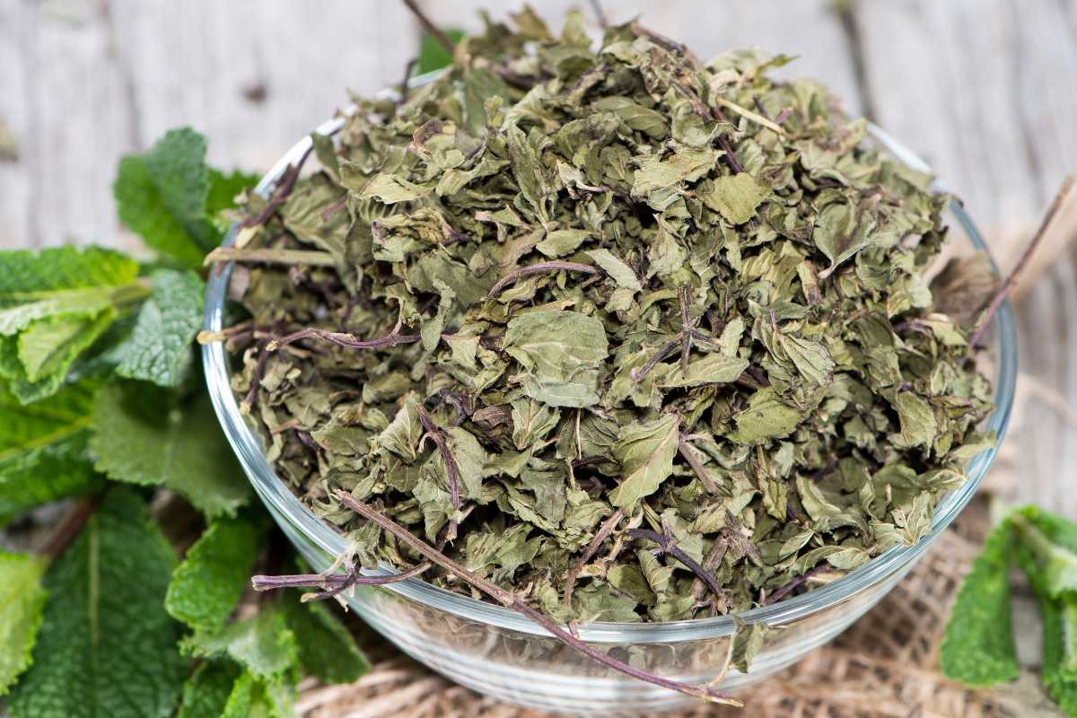 A clear glass bowl filled with dried mint leaves rests on a rustic wooden surface. 