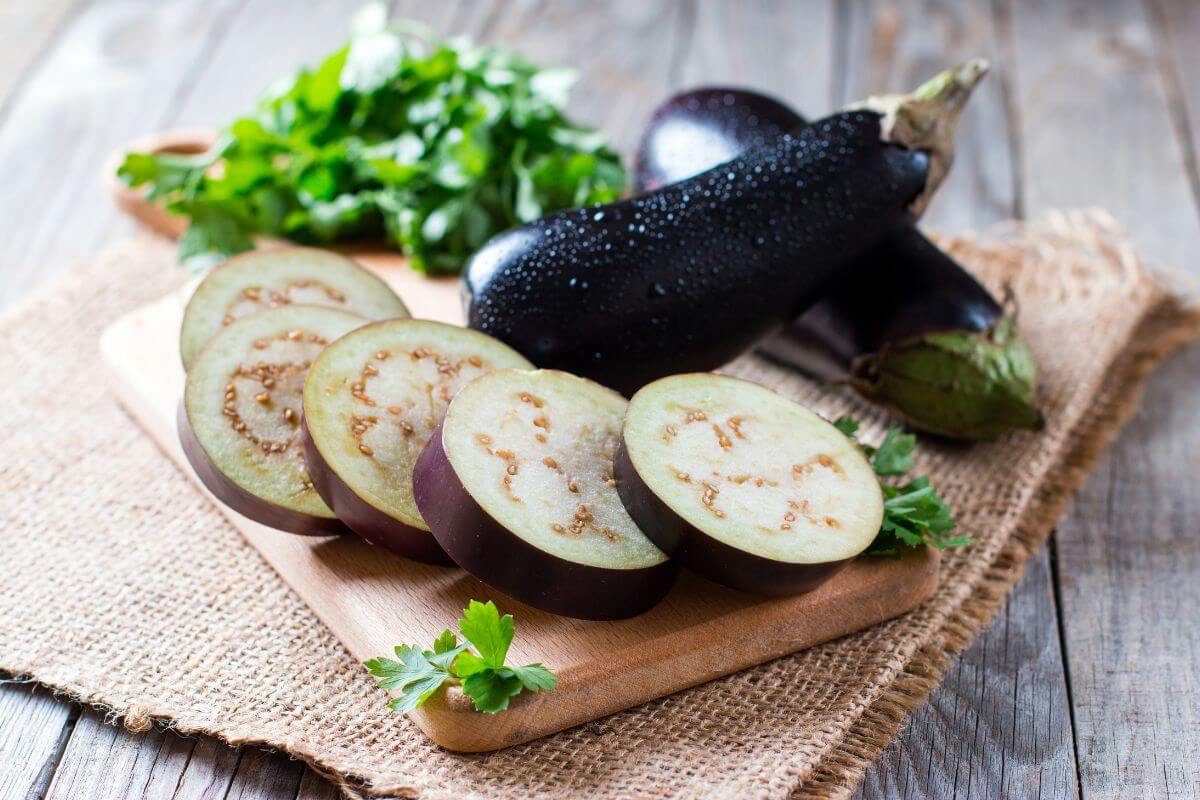 Sliced eggplant on a wooden cutting board, with whole eggplants and fresh parsley in the background, set on a burlap cloth atop a wooden table.