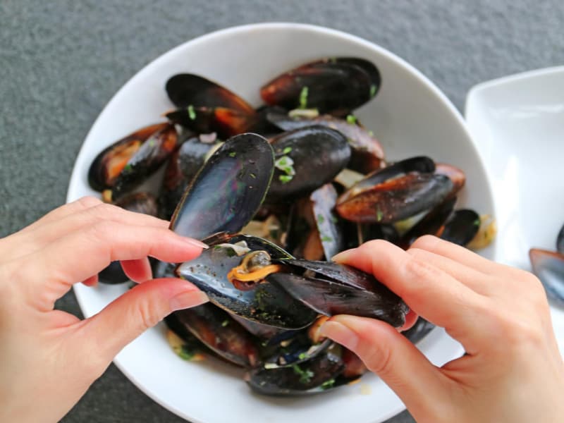 Close-up of hands opening a mussel over a bowl of cooked mussels.